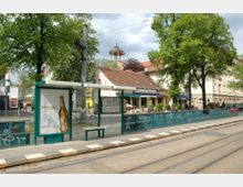 Straßenbahnhaltestelle mit Wartehäuschen und Werbeposter im Vordergrund, im Hintergrund ein Restaurant mit der Beschriftung „Ristorante Leonardo“, umgeben von Bäumen und einem Turm mit Kuppeldach.