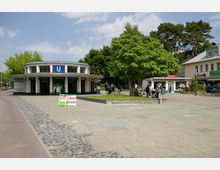 Ein kleiner U-Bahnhof mit rundem Eingang und blauem "U"-Zeichen im Zentrum eines gepflasterten Platzes. Links ein Weg, rechts mehrere Geschäfte sowie ein Baum und Fahrräder im Hintergrund.
