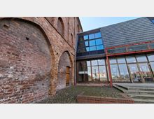 Innenhof mit historischer Backsteinfassade und großen gemauerten Rundbögen, daneben ein moderner Glas-Anbau mit großen Fensterflächen. Kopfsteinpflaster, eine kleine Holz­tür und eine Treppe führen zu einem verglasten Eingangsbereich unter einem schiefergedeckten Dach.