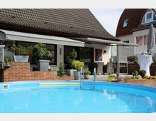 Wohnhaus mit einer Terrasse und einem runden, blauen Außenpool im Vordergrund. Die Terrasse ist mit Pflanzen in Töpfen, Gartenmöbeln und einem Grill ausgestattet, während im Hintergrund ein weißes Haus mit großen Fenstern zu sehen ist.