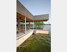 Modernes Gebäude mit Holzfassade, direkt über dem Wasser eines Sees gebaut. Große Glasfronten, ein Zugang über eine Treppe ins Wasser und eine umlaufende Terrasse mit Geländer. Im Hintergrund sind Bäume und der Himmel sichtbar.