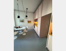 Minimalistisch eingerichtete Küche mit pastellfarbenen Einbauschränken und grauem Boden. Im Zentrum steht ein runder Esstisch mit weißen Stühlen, an der Wand hängen zwei Bilder und eine Uhr, von der Decke hängen mehrere Lampen mit schwarzen Kabeln.