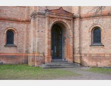 Backsteinfassade eines Gebäudes mit einem hohen Rundbogenportal, verziert mit Ornamenten, flankiert von zwei Rundbogenfenstern mit Gitterverglasung. Vor dem Eingang befinden sich steinerne Stufen, umgeben von einem kleinen Rasenstück.