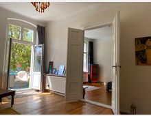 Helles Wohnzimmer mit Holzboden und weißen Wänden, ausgestattet mit einer großen Doppeltür, die in einen angrenzenden Raum führt, sowie einer offenen Balkontür mit Blick auf eine grüne Außenterrasse. Im Vordergrund befinden sich Möbel und ein Heizkörper mit darauf aufgestellten Bilderrahmen.