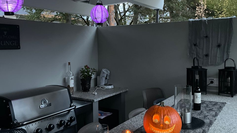 Überdachte Outdoor-Küche mit grauen Arbeitsflächen, einem Edelstahlgrill, lila Laternen an der Decke und dekorativen Elementen wie einem beleuchteten Kürbis und Kerzen auf dem Tisch. Im Hintergrund befinden sich graue Wände und Bäume außerhalb der offenen Seiten.