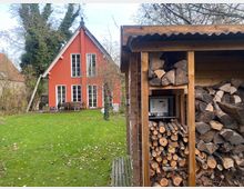 Ein rotes Einfamilienhaus mit Satteldach und großen weißen Fenstern steht in einem Garten mit Rasen und laubabwerfenden Bäumen. Im Vordergrund befindet sich ein Holzlager mit gestapeltem Brennholz und einem kleinen Schaltkasten.