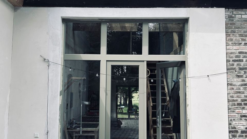 Innenhofansicht eines Hauses mit rotem Ziegeldach und Dachfenster sowie großer, bodentiefer Glasfront mit Rahmen und Oberlicht. Vor dem Eingang liegen Kopfsteinpflaster, mehrere Blumentöpfe und eine Gießkanne; seitlich sind ein kleiner Tisch und eine Ziegelwand zu sehen.