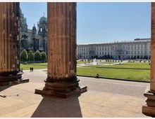 Blick durch massive Steinsäulen auf einen offenen Platz mit Rasenflächen, einem Springbrunnen und Spazierwegen. Im Hintergrund sind der Berliner Dom mit seiner Kuppel sowie ein historisches Gebäude mit klassizistischer Fassade zu sehen.