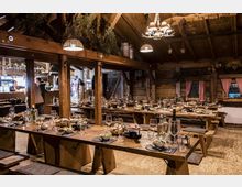 Rustikaler Speiseraum in einer Holzhütte mit sichtbaren Holzbalken und warmen Hängelampen, ausgestattet mit langen Holztischen und Bänken. Die Tische sind festlich gedeckt mit Gläsern, Geschirr und Speisen, neben Fenstern mit rosa Vorhängen und Holzvertäfelung.