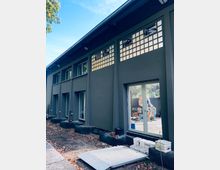 Ein modernes Gebäude mit dunkelgrauer Fassade und großen Fenstern in einer Reihe. Die oberen Fenster bestehen aus Glasbausteinen, und im Vordergrund befinden sich Baustellenmaterialien sowie eine Rampe.