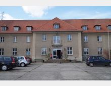 Ein mehrstöckiges Gebäude mit beigem Putz und einem roten Satteldach, in der Mitte ein Eingangsbereich mit einer kleinen Treppe und einem Balkon darüber. Auf dem Vorplatz stehen Autos geparkt, und einige Personen befinden sich im Eingangsbereich.