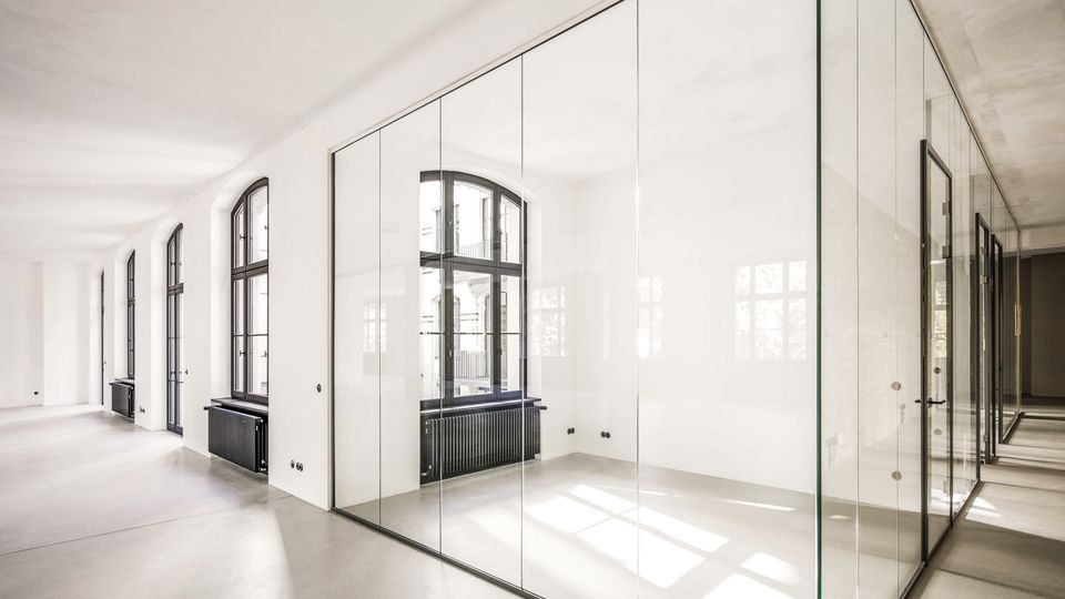 Heller, minimalistischer Innenraum eines modernen Büro- oder Ateliergebäudes mit poliertem Betonboden, weißen Wänden und hohen, bogenförmigen Fenstern entlang der linken Seite. In der Mitte steht ein gläserner Kubus als abgetrennter Raum, dessen transparente Wände Licht und Fensterreflexionen zeigen.