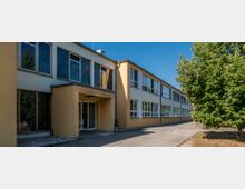 Zweistöckiges, modernes Gebäude mit gelber Fassade und großen Fenstern, die teils mit Musterverglasung versehen sind. Ein gepflasterter Gehweg führt zum Eingang, und an der Seite steht ein großer Baum.