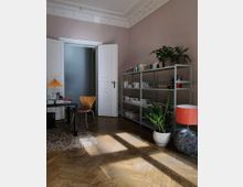 Ein stilvoll eingerichtetes Zimmer mit Parkettboden, hohen Decken und verzierten Stuckleisten. Zu sehen sind ein offener weißer Türrahmen, ein Regal mit Büchern und Pflanzen, ein Schreibtisch mit einer Lampe sowie ein gemütlicher Teppich auf dem Boden.