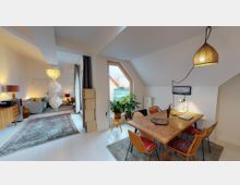 Wohnbereich mit offener Raumaufteilung, der ein Esszimmer mit einem Holztisch und bunten Stühlen sowie ein Wohnzimmer mit einem Sofa und einer Stehlampe umfasst. Große Fenster mit Dachschrägen lassen viel Licht herein, und Pflanzen sowie dekorative Details prägen den gemütlichen Stil der Einrichtung.