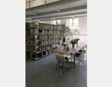 Heller, moderner Innenraum mit einer großen Bücherwand, einem Holztisch, weißen Stühlen und dekorativen Pflanzen. Große Fenster lassen natürliches Licht herein, und eine offene Metallkonstruktion prägt die Decke.