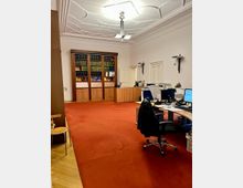 Ein Büro mit rotem Teppich, mehreren modernen Schreibtischen und Bürostühlen. Im Hintergrund steht ein großer Holzschrank mit Büchern, und die Decke hat dekorative Stuckdetails.