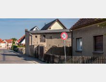 Straßenszene in einer Wohnsiedlung mit mehreren Häusern mit Ziegeldächern und einfachem Putz. Im Vordergrund steht ein Verkehrsschild mit einer Geschwindigkeitsbegrenzung von 50 km/h, und ein niedriger Holzzaun grenzt die Grundstücke ab.