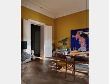 Wohnzimmer mit gelber Wand, Holztisch und hölzernen Stühlen, teilweise mit Polstern. An der Wand hängt ein großes, farbiges Kunstwerk, und auf dem Tisch befinden sich Bücher, Kerzenleuchter und Dekorationsgegenstände. Der Boden besteht aus Fischgrätenparkett, und durch geöffnete, weiße Doppeltüren fällt Tageslicht in den Raum.