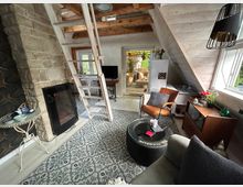 Gemütlicher Wohnraum in einem Dachgeschoss mit hellem Holz und Holzbalkendecke. Zu sehen sind ein gemauerter Kamin mit Elektrofeuer, eine steile Holzleiter, ein Teppich mit Muster, eine Sitzecke mit Ledersessel und Couchtisch sowie eine Tür zu einer Terrasse mit Glasüberdachung.