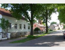Straßenszene in einem Wohngebiet mit Einfamilienhäusern, gepflastertem Gehweg und einer Allee aus großen Bäumen, die Schatten auf die Straße werfen. Im Hintergrund sind weitere Häuser sowie ein hölzerner Turm sichtbar.