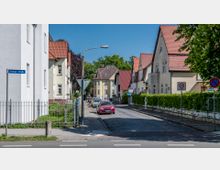 Straßenszene in einem Wohngebiet mit gepflasterter Fahrbahn, flankiert von Einfamilienhäusern mit roten Ziegeldächern und hellem Putz. Die Straße ist mit Tempo-30-Schildern markiert, und ein Straßenschild mit der Aufschrift "Berliner Straße" ist links zu sehen.