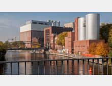 Industriegebäude mit roten Backsteinfassaden und modernen silbernen Silotürmen an einem Flussufer, umgeben von herbstlich gefärbten Bäumen. Im Vordergrund verläuft ein Geländer, und eine Fußgängerbrücke führt über den Fluss.