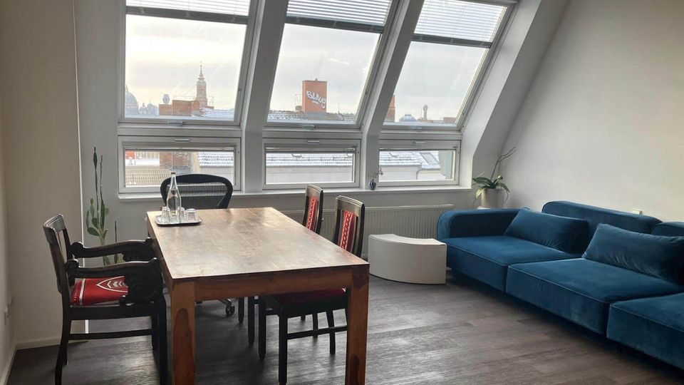 Attic-style living and dining room with a sloped ceiling and large angled skylight windows overlooking city rooftops. A wooden dining table with chairs sits on dark flooring, with a blue sectional sofa and small white side tables along the wall.