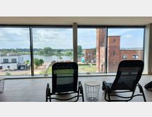 Blick durch große Panoramafenster auf eine Flusslandschaft mit einem roten Backsteingebäude, moderner Architektur und Brücke im Hintergrund. Im Vordergrund stehen zwei Liegestühle und ein kleiner Beistelltisch in einem Innenraum mit Fliesenboden.