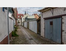 Schmale, gepflasterte Gasse mit niedrigen Gebäuden und Garagen aus Metall und Ziegeln zu beiden Seiten; am Ende der Gasse sind Wohnhäuser mit roten und hellen Fassaden sowie ein Kirchturm sichtbar.
