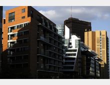 Städtische Skyline mit modernen Gebäuden aus Glas, Stahl und Backstein; links ein Wohngebäude mit Balkonen, rechts Bürokomplexe mit markanten Fassaden und vorgelagerten Sonnenschutzstrukturen. Im Hintergrund ein hoher Turm mit dunkler Fassade und Antenne.