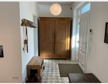 Flur mit heller Wandgestaltung und gemustertem Fliesenboden. Zu sehen sind ein großer Holzschrank, eine Garderobe mit Haken, ein Holzbank mit Schuhablage sowie eine weiße Eingangstür mit Glaseinsätzen und Oberlichtern.