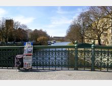 Eine Brücke mit dekorativem Metallgeländer über einen Fluss in einer urbanen Umgebung. Im Vordergrund steht ein Zeitungsständer, im Hintergrund sind Bäume, Wohnhäuser und Boote auf dem Wasser zu sehen.