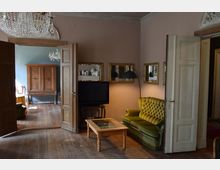 Wohnzimmer in einem historischen Gebäude mit Holzböden und cremefarbenen Wänden. Im Vordergrund ein grünes Sofa, ein Couchtisch und ein Fernseher, im Hintergrund ein angrenzender Raum mit einem Holzschrank und Kronleuchtern.