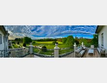 Panoramablick von einer Terrasse mit elegantem Geländer auf eine weitläufige Parklandschaft mit Rasenflächen, Bäumen und einem klaren Himmel mit Wolkenformationen. Auf der Terrasse stehen Sitzmöbel und Tische, die zum Verweilen einladen.