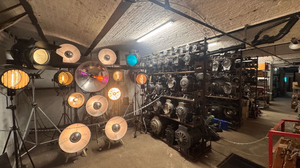 Raum mit industriellem Charakter und gewölbter Backsteindecke, gefüllt mit alten Film- und Bühnenscheinwerfern auf Ständern sowie Regalen, die mit weiteren Scheinwerfern und Equipment bestückt sind. Der Raum ist spärlich beleuchtet, wobei einige Scheinwerfer eingeschaltet sind und warmes Licht abstrahlen.