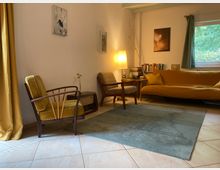 Wohnzimmer mit Retro-Möbeln: Zwei Sessel, einer in Senfgelb, der andere in Grau, stehen neben einem kleinen Tisch mit Lampe. Ein gelbes Sofa, ein Regal mit Büchern und ein großes Fenster mit grünem Ausblick befinden sich im Hintergrund. Ein blauer Teppich bedeckt den gefliesten Boden.