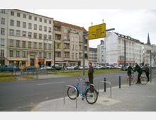 Straßenszene in einem städtischen Gebiet mit mehrstöckigen Wohn- und Geschäftsgebäuden im Hintergrund. Im Vordergrund ein Radständer, geparkte Fahrräder und Passanten auf dem Bürgersteig, während Autos auf der Straße vorbeifahren; ein Verkehrsschild weist auf die Richtungen "Friedrichshain" und "Mitte" hin.