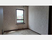 Ein kleiner, leerer Raum mit verblasster, gemusterter Tapete und einem rechteckigen Fenster, das Tageslicht hereinlässt. Der Boden ist abgenutzt, und das Fenster bietet einen Blick auf ein Dach mit roten Ziegeln und Bäume im Hintergrund.