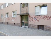 Mehrfamilienhaus mit beigefarbener Fassade und Klinkersockel, mehreren Fenstern mit Rollläden und einem verglasten Hauseingang. An der Wand sind rote und weiße Graffitis zu sehen, und der Gehweg ist mit Herbstlaub bedeckt.