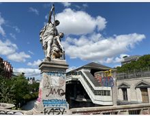 Statue eines Mannes und eines Jungen mit Waffe auf einem Sockel, der mit Graffiti bedeckt ist, vor einer Brücke und einem Gebäude mit einem erhöhten Treppenaufgang. Im Hintergrund sichtbar sind ein Kanal, Bäume und moderne Gebäude unter einem blauen Himmel mit vereinzelten Wolken.
