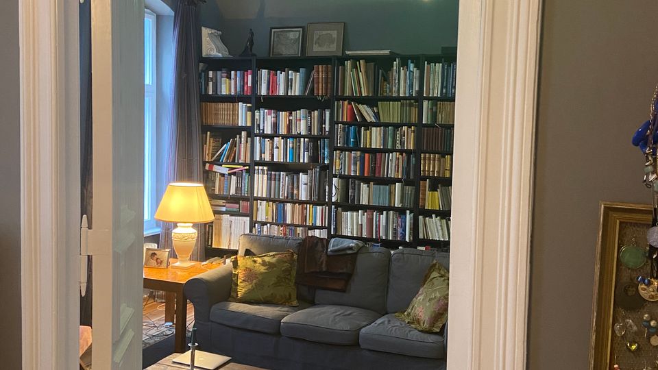 Blick durch eine Tür in ein Wohnzimmer mit hoher Stuckdecke, grünem Hängelicht und einer wandfüllenden Bücherregalwand. Im Raum stehen ein graues Sofa, ein niedriger Holztisch auf einem großen Teppich sowie ein Beistelltisch mit Lampe neben einem Fenster.