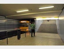 U-Bahn-Station mit gelbem Zug, der an einem Gleis vorbeifährt, einer Treppe, die zu einem Ausgang mit dem Schild "Ausgang" und "Olsoer Straße" führt, sowie einer Person, die sich vom Gleisbereich entfernt. Die Wände sind mit grauen und blauen Fliesen verkleidet, die Decke hat einfache Leuchtstofflampen.