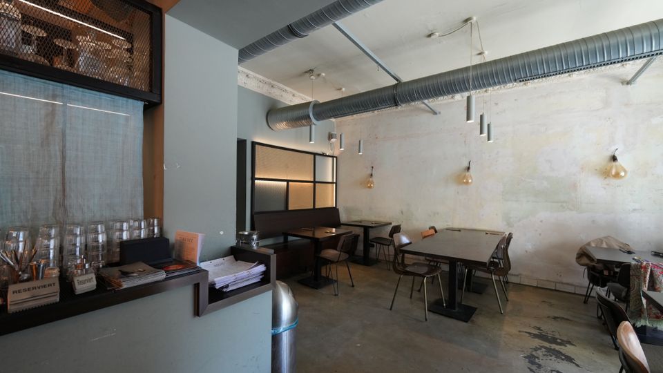 Innenraum eines industriell gestalteten Cafés oder Restaurants mit hohen Decken, sichtbaren Lüftungsrohren und hängenden Leuchten. Links eine Theke mit gestapelten Gläsern, rechts mehrere einfache Tische und Stühle vor einer hellen, roh verputzten Wand.