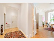 Heller Flur in einer Altbauwohnung mit weißen Wänden, Stuckdecke, hohen Türen und Parkettboden mit gemusterten Teppichen. Rechts öffnet sich der Blick in ein Zimmer mit großen Fenstern, Tisch und Stühlen sowie Zimmerpflanzen.