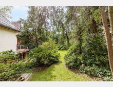 Ein gepflegter Garten mit einer Rasenfläche, umgeben von dichten Sträuchern und hohen Nadelbäumen, angrenzend an ein Wohnhaus mit hellen Fassaden und einem Balkon mit Metallgeländer. Im Hintergrund sind weitere grüne Pflanzen und Blumenbeete zu erkennen.