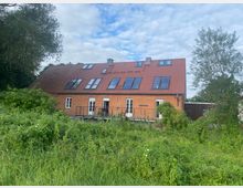 Ein modernes, zweistöckiges Gebäude aus rotem Backstein mit einem roten Satteldach und mehreren großen Dachfenstern. Im Vordergrund befindet sich eine dicht bewachsene Wiesenfläche, umgeben von Bäumen und Sträuchern.