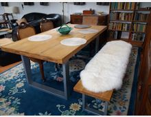 Esszimmer mit einem rechteckigen Holztisch und dazu passenden Stühlen mit Geflecht und einer Bank mit einem weißen Fellpolster. Im Hintergrund befinden sich Bücherregale, ein Teppich mit floralen Mustern liegt auf dem Boden, und Loungesessel sind sichtbar.