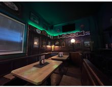 Gemütlicher Pub-Innenraum mit dunkler Holzvertäfelung, Eckbänken und mehreren Holztischen, auf denen Speisekarten stehen. An den Wänden hängen gerahmte Bilder und ein Schild „Off to an Rboist“, dazu gedämpfte Lampen und grüne indirekte Beleuchtung unter der Decke.