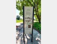 Eine Informationsstele im Freien mit der Überschrift „Park und Schlösschen Monplaisir“, die historischen Text und Illustrationen zeigt, darunter eine Zeichnung des Schlosses. Im Hintergrund sind ein gepflasterter Weg, Wiese und Bäume zu sehen, die für eine parkähnliche Umgebung typisch sind.